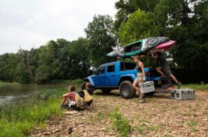A blue truck is parked by a riverbank with two kayaks on its roof. Two people unload gray LINE-X coolers while two kids sit nearby, gazing at the river.