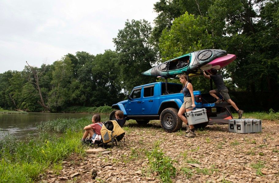 A blue truck is parked by a riverbank with two kayaks on its roof. Two people unload gray LINE-X coolers while two kids sit nearby, gazing at the river.