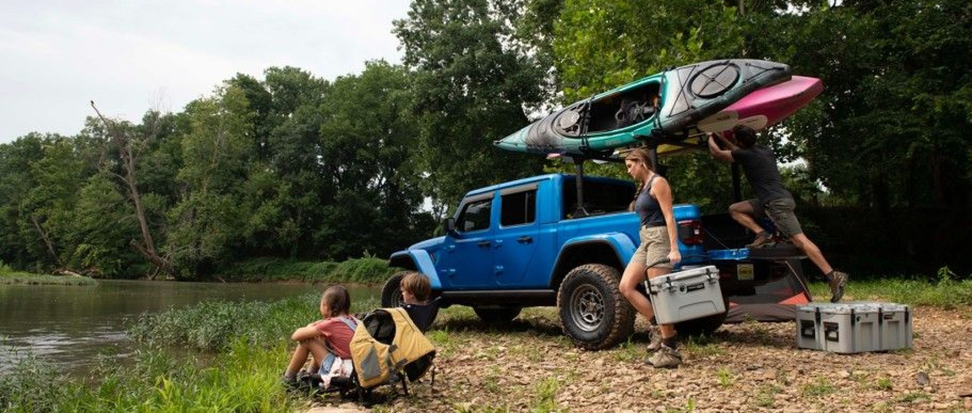 A blue truck is parked by a riverbank with two kayaks on its roof. Two people unload gray LINE-X coolers while two kids sit nearby, gazing at the river.
